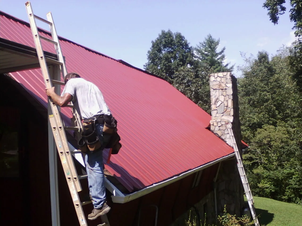 Roofer on a ladder beside a red metal roof and stone chimney — Soffit & Fascia Repair by Roofing Brothers in East Vandergrift